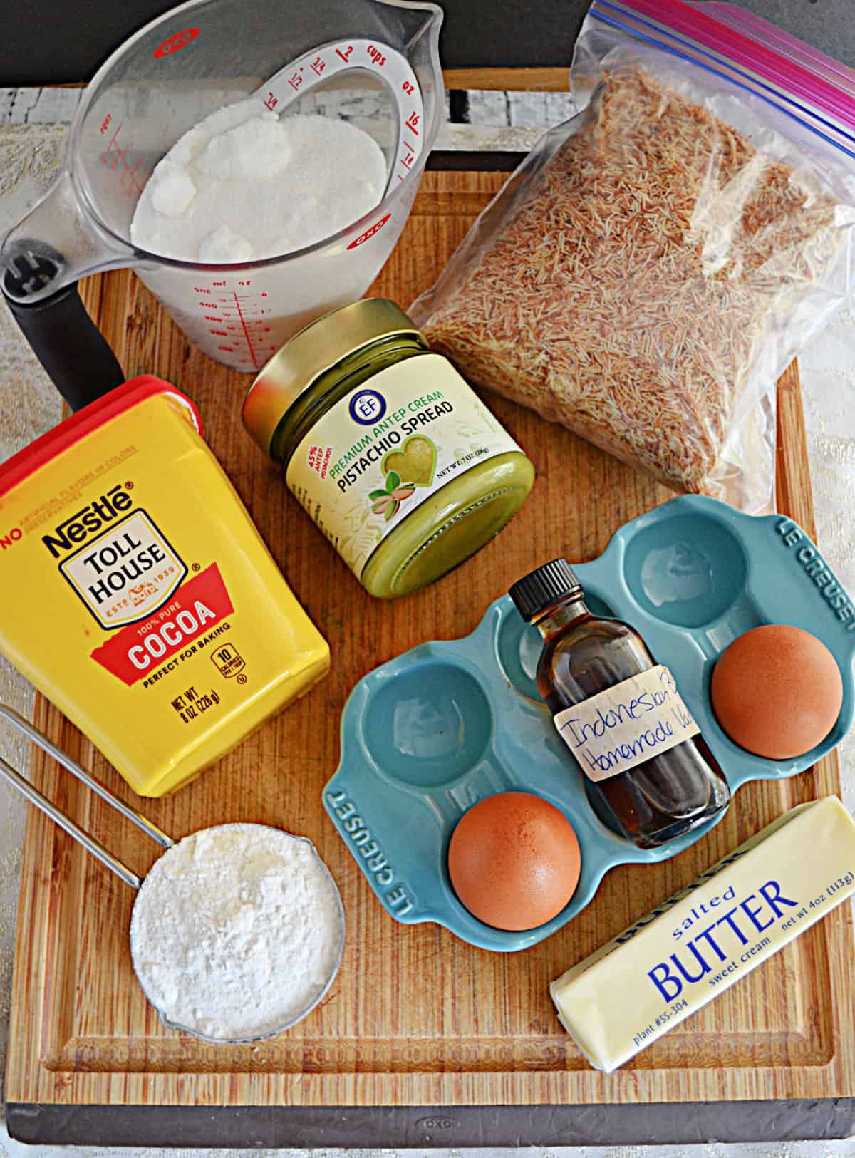 A cutting board with flour, cocoa powder, pistachio cream, shredded phyllo dough, eggs, and vanilla on it.