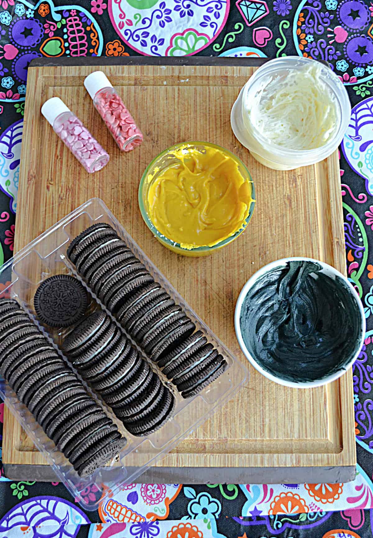 A cutting board with three colors of frosting/chocolate, two containers of sprinkles, and a sleeve of chocolate cookies.