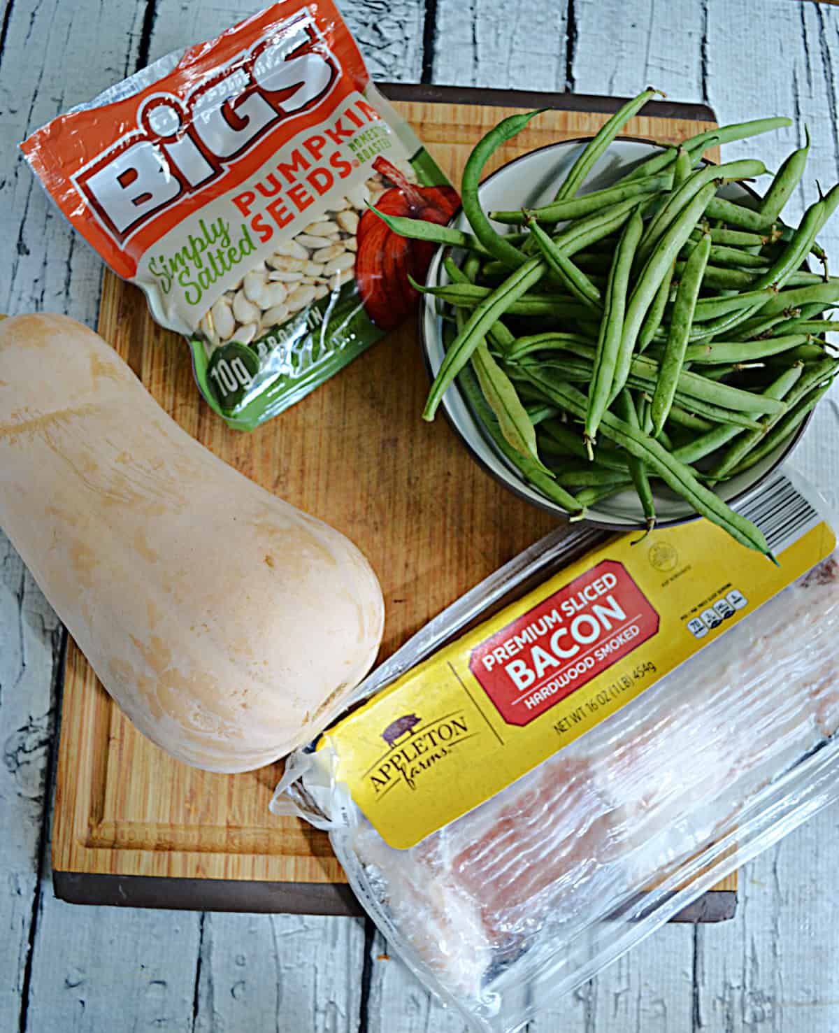 A cutting board with a butternut squash, a package of bacon, a bowl of green beans, and a package of pumpkin seeds on it.