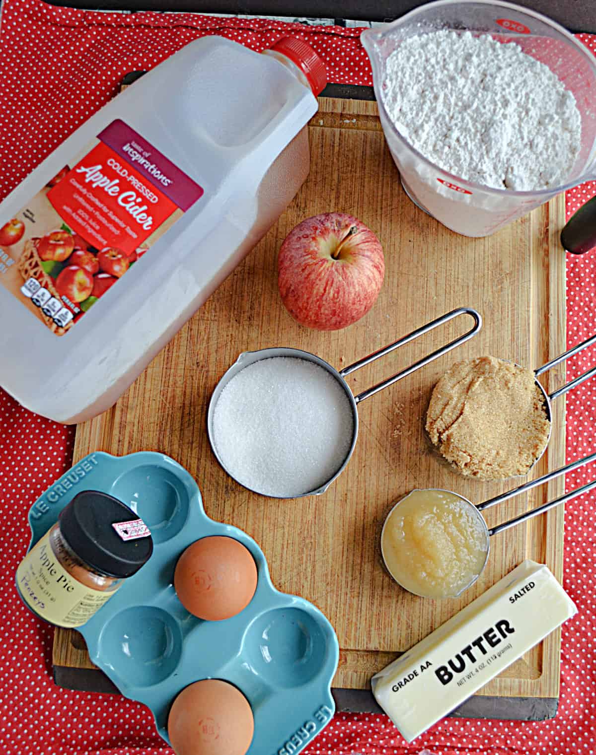 A wooden cutting board with a carton of apple cider, an apple, a cup of flour, a cup of sugar, eggs, a stick of butter, and spice jars on it.