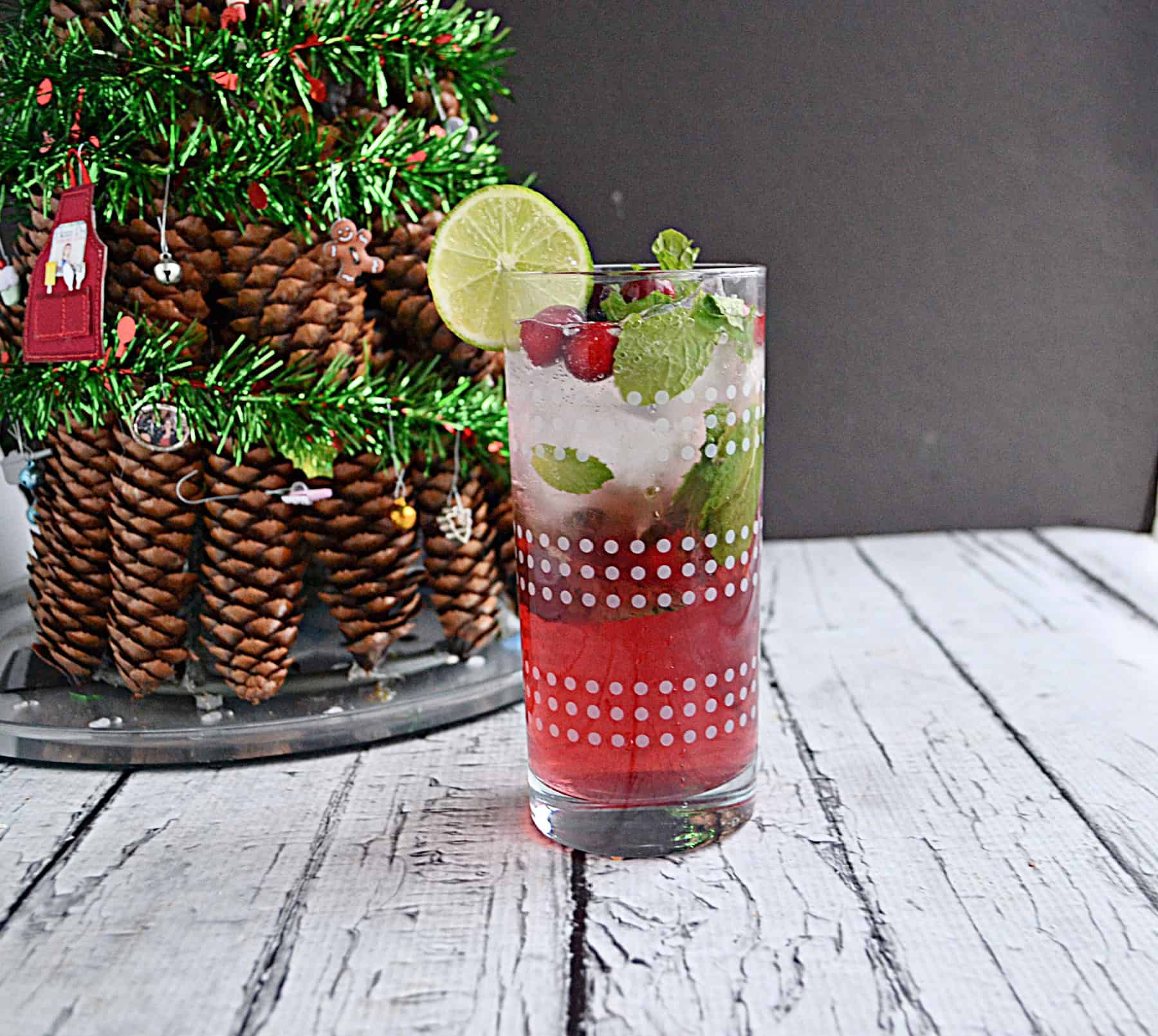 A glass of mojito with a lime slice and mint leaves by a Christmas tree.