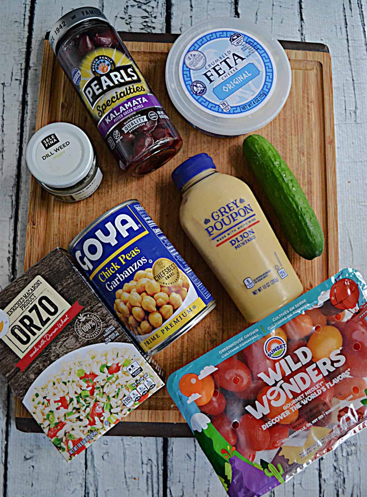 A cutting board with a box of orzo, a can of chickpeas, a container of tomatoes, olives, a cucumber, mustard, and feta cheese.
