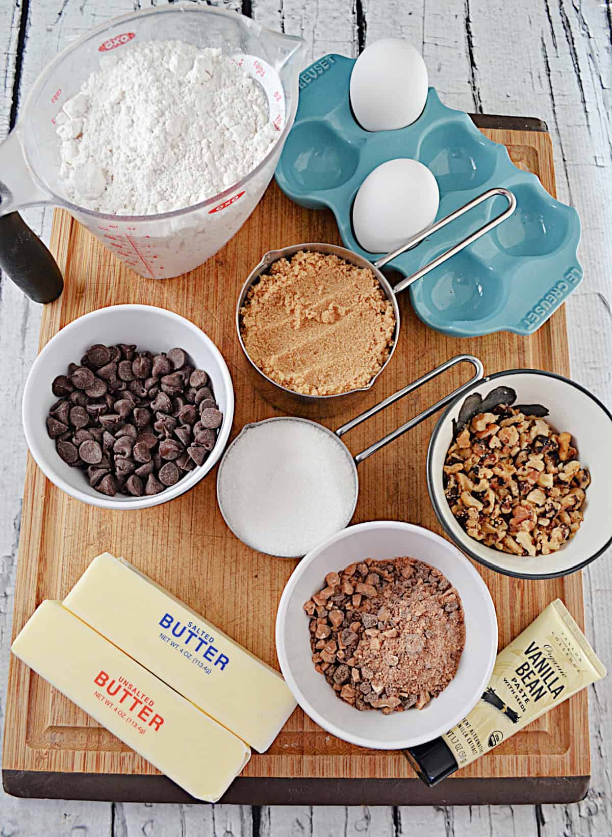 A cutting board with a cup of flour, a cup of chocolate chips, a cup of nuts, cups of sugar, two sticks of butter, and eggs on it.