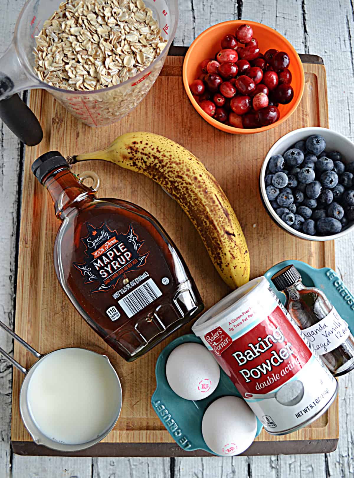A wooden cutting board with a cup of oats, bowls of fruit, a banana, a jar of maple syrup, eggs, and sugar.
