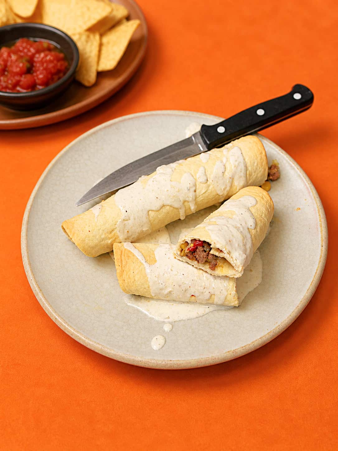 A plate with two taquitos on i and a knife on the plate, with a plate of chips and salsa in the background.