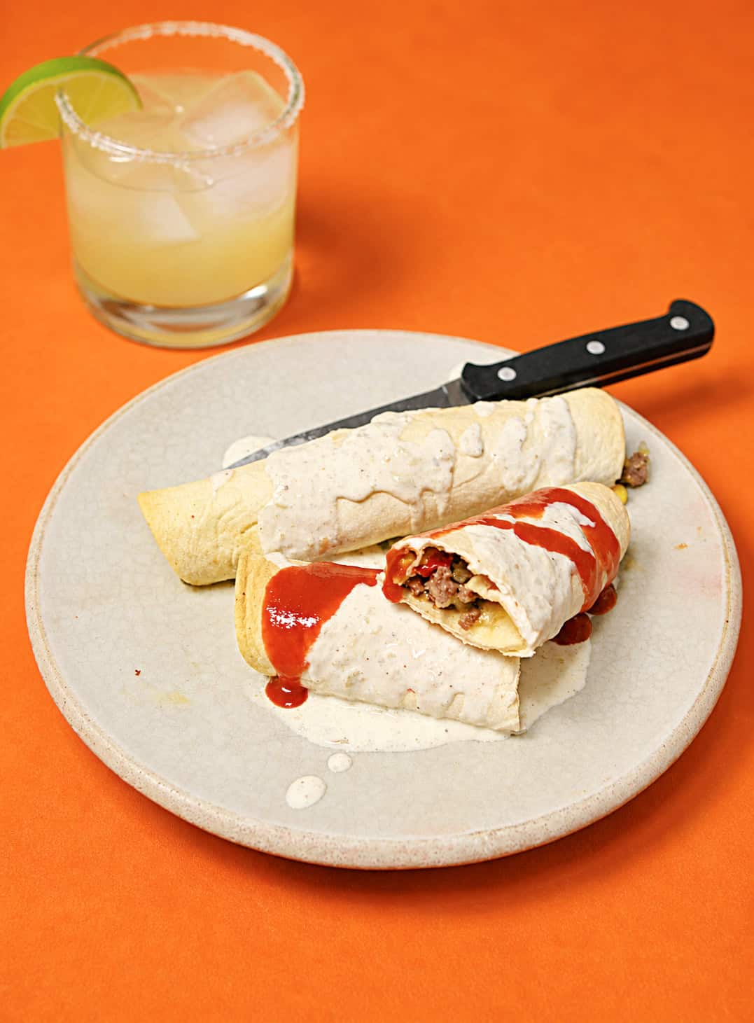 A plate of taquitos with white sauce and a margarita behind the plate.