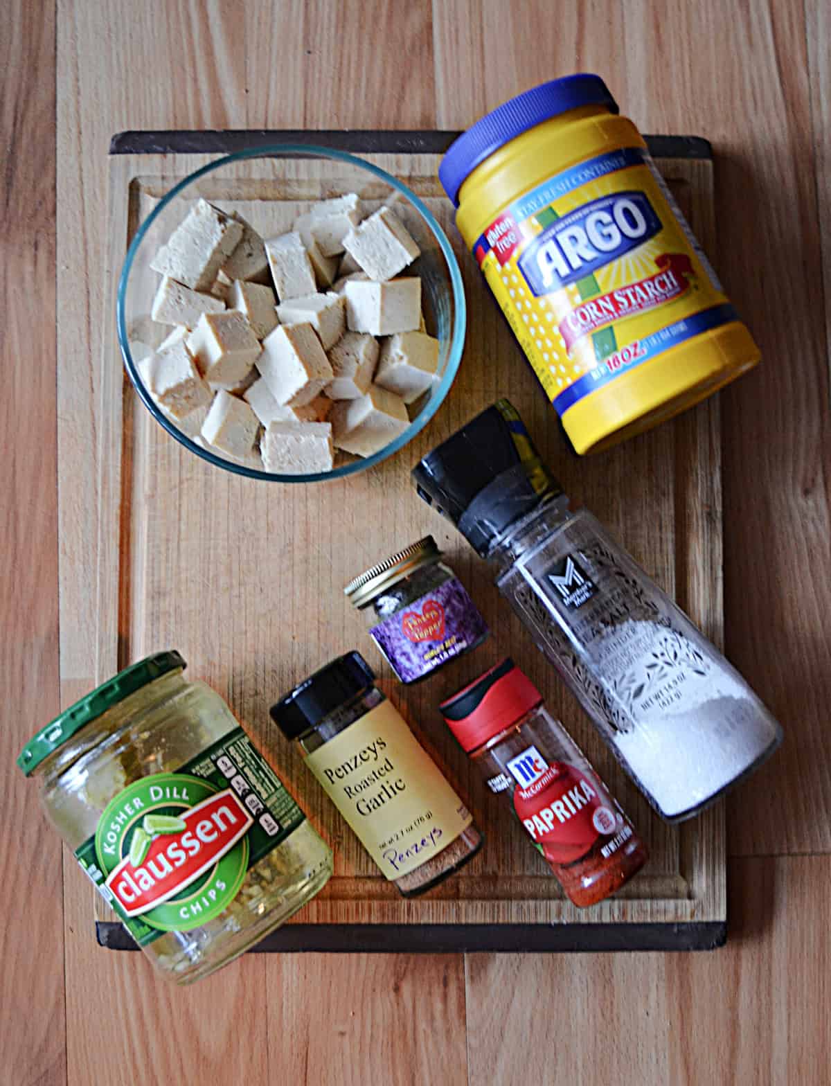A wooden cutting board with a bowl of tofu, a container of corn starch, a jar of pickles, and spices.