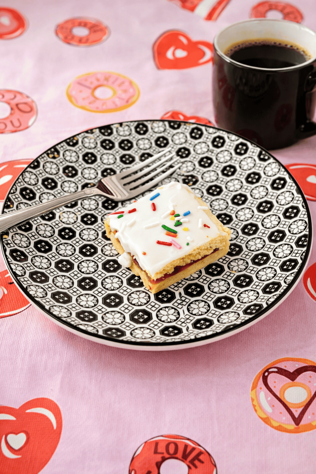 A plate with a cookie bar with white icing and sprinkles on it, a fork, and a mug of coffee.