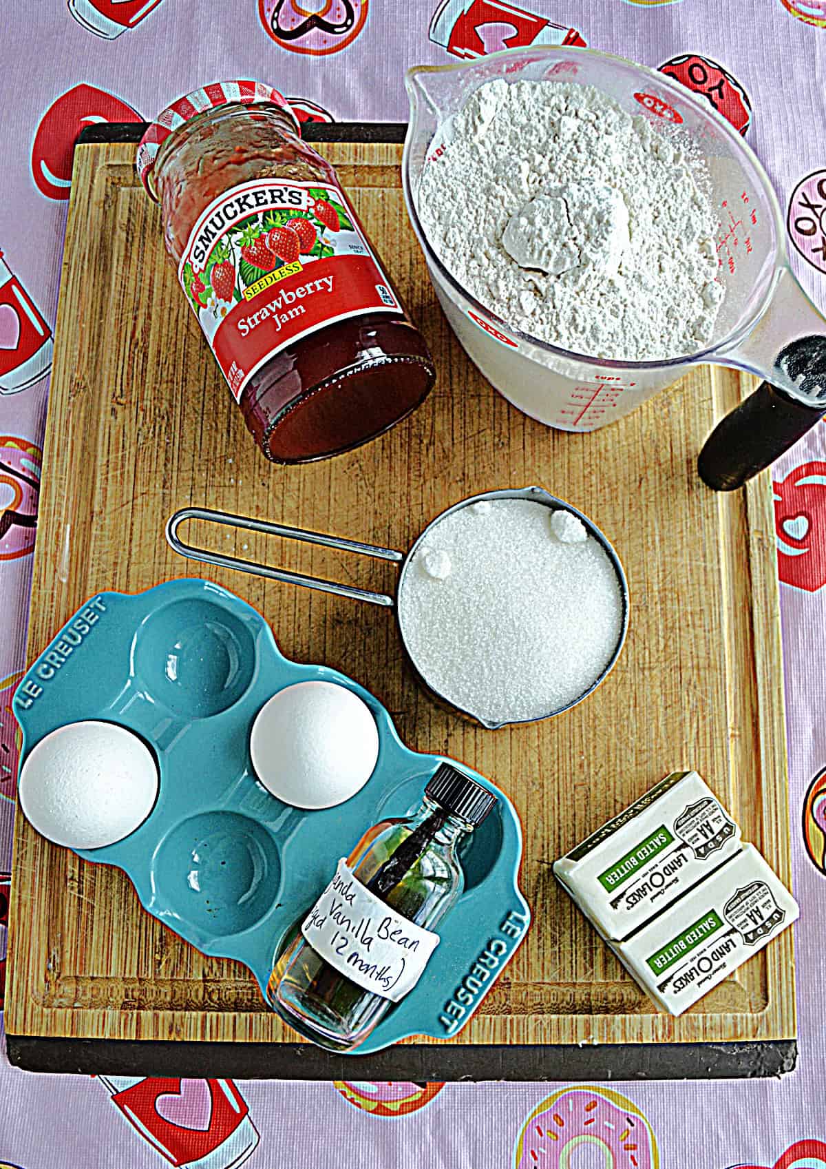 A wooden cutting board with a cup of flour, a cup of sugar, butter, eggs, vanilla, and a jam jar on it.