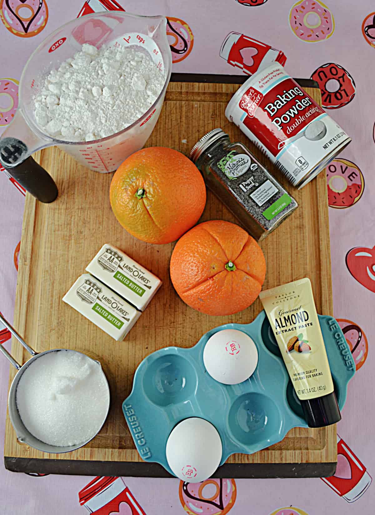 A wooden cutting board with oranges, a cup of flour, a cup of sugar, butter, poppyseeds, eggs, and almond.