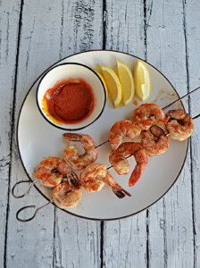 A plate with grilled shrimp skewers, lemon wedges, and a bowl of cocktail sauce.