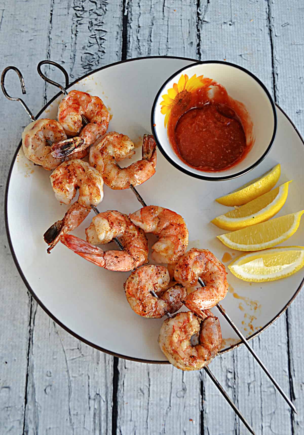 A plate with grilled shrimp on skewers, lemon wedges, and a bowl of cocktail sauce.