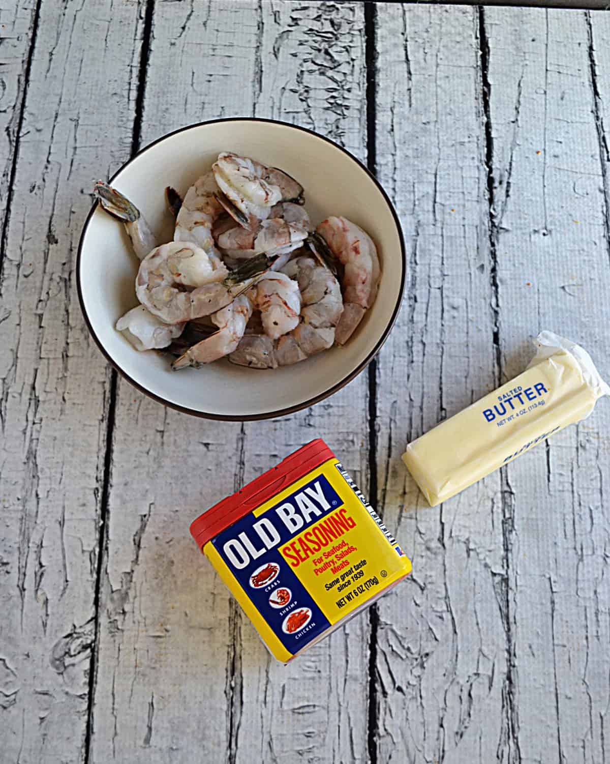 A bowl of raw shrimp, a container of Old Bay Seasoning, and a stick of butter.