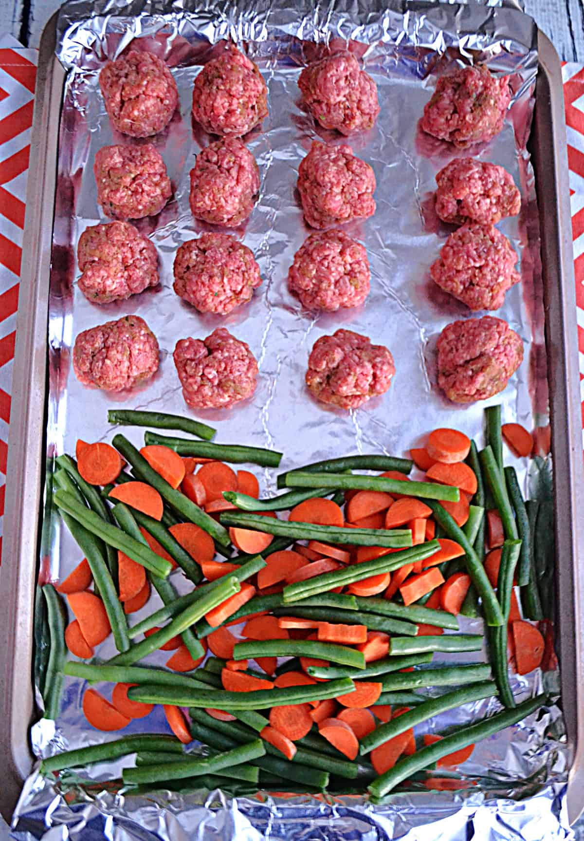 A sheet pan with uncooked meatballs, carrots, and green beans on it.