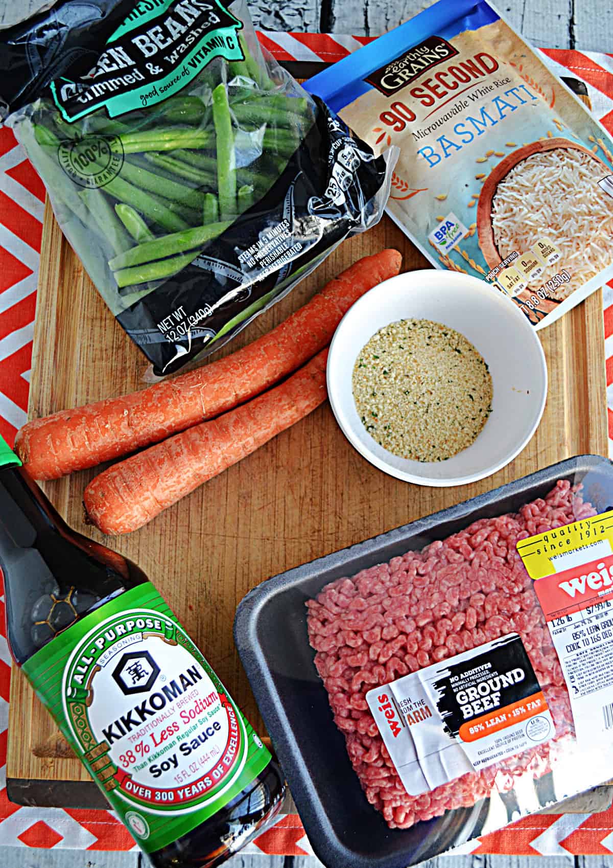 A cutting board with green beans, rice, carrots, soy sauce, ground beef, and breadcrumbs on it.