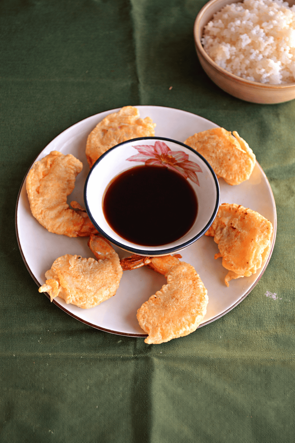 A plate with golden brown shrimp surrounding a bowl of dipping sauce and a bowl of rice in the background.