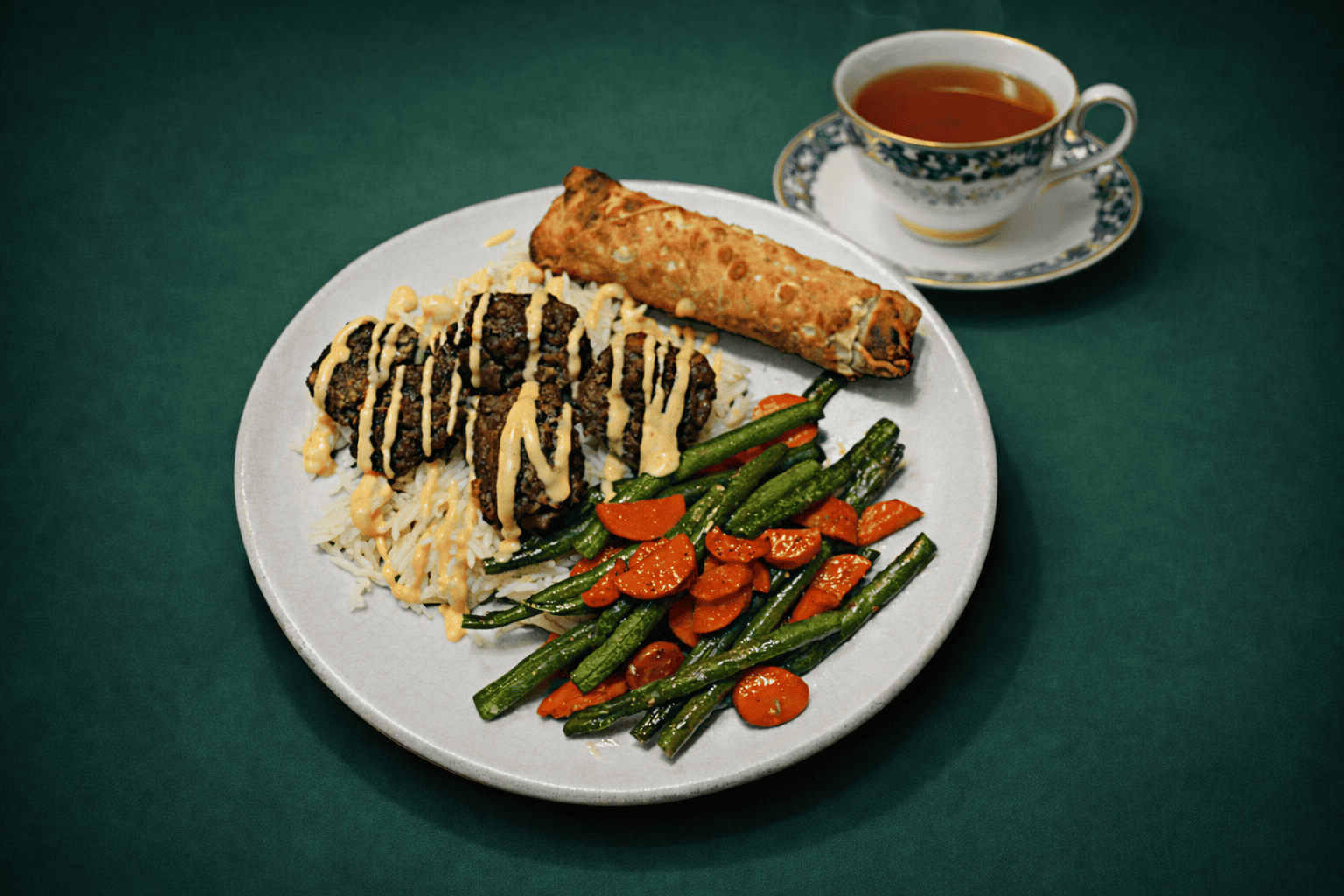 A plate with meatballs on rice with sauce on top, a pile of veggies, and an eggroll and a cup of hot tea in the background.