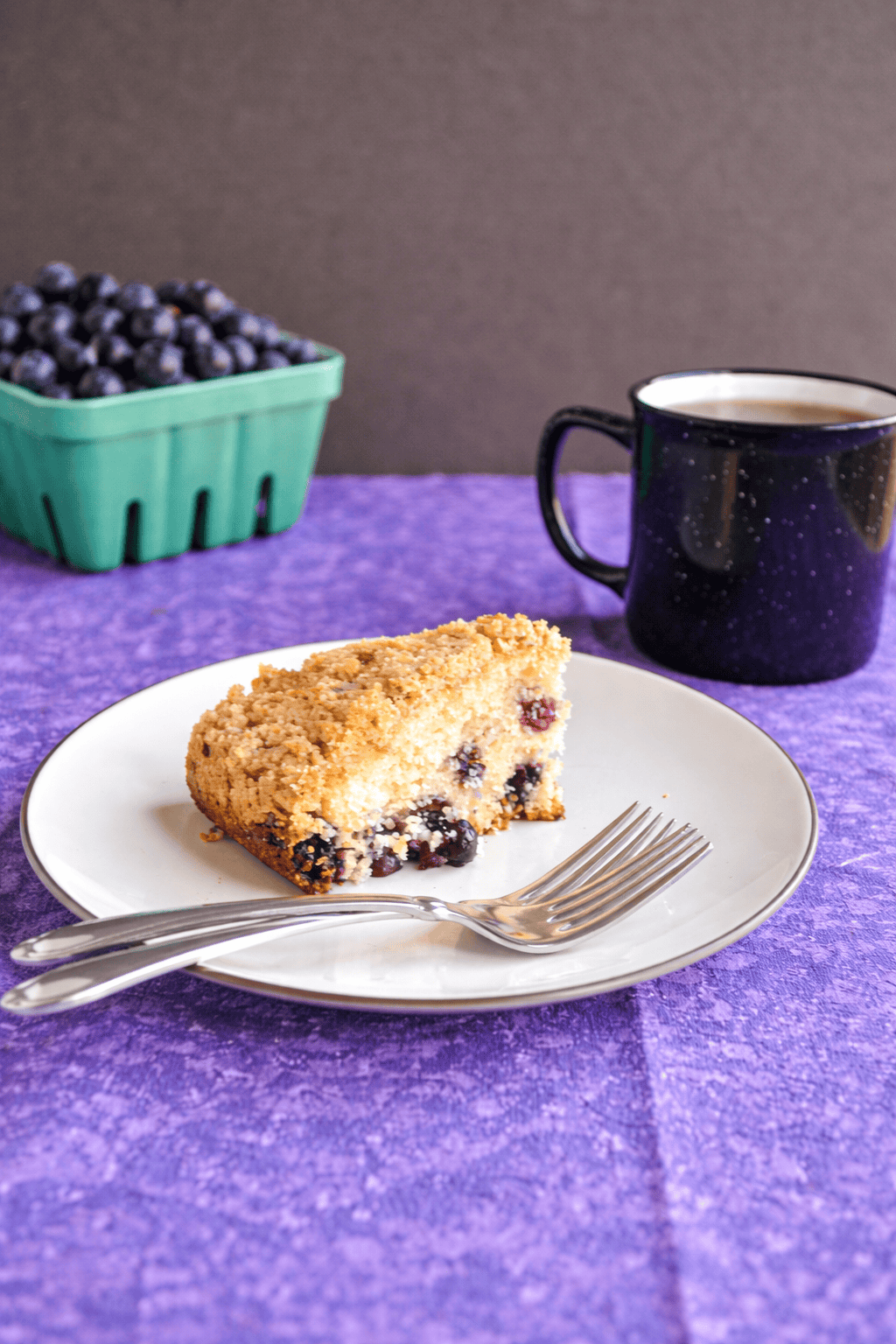 A plate with a slice of coffee cake on it, a mug of coffee behind the plate, and a container of blueberries.