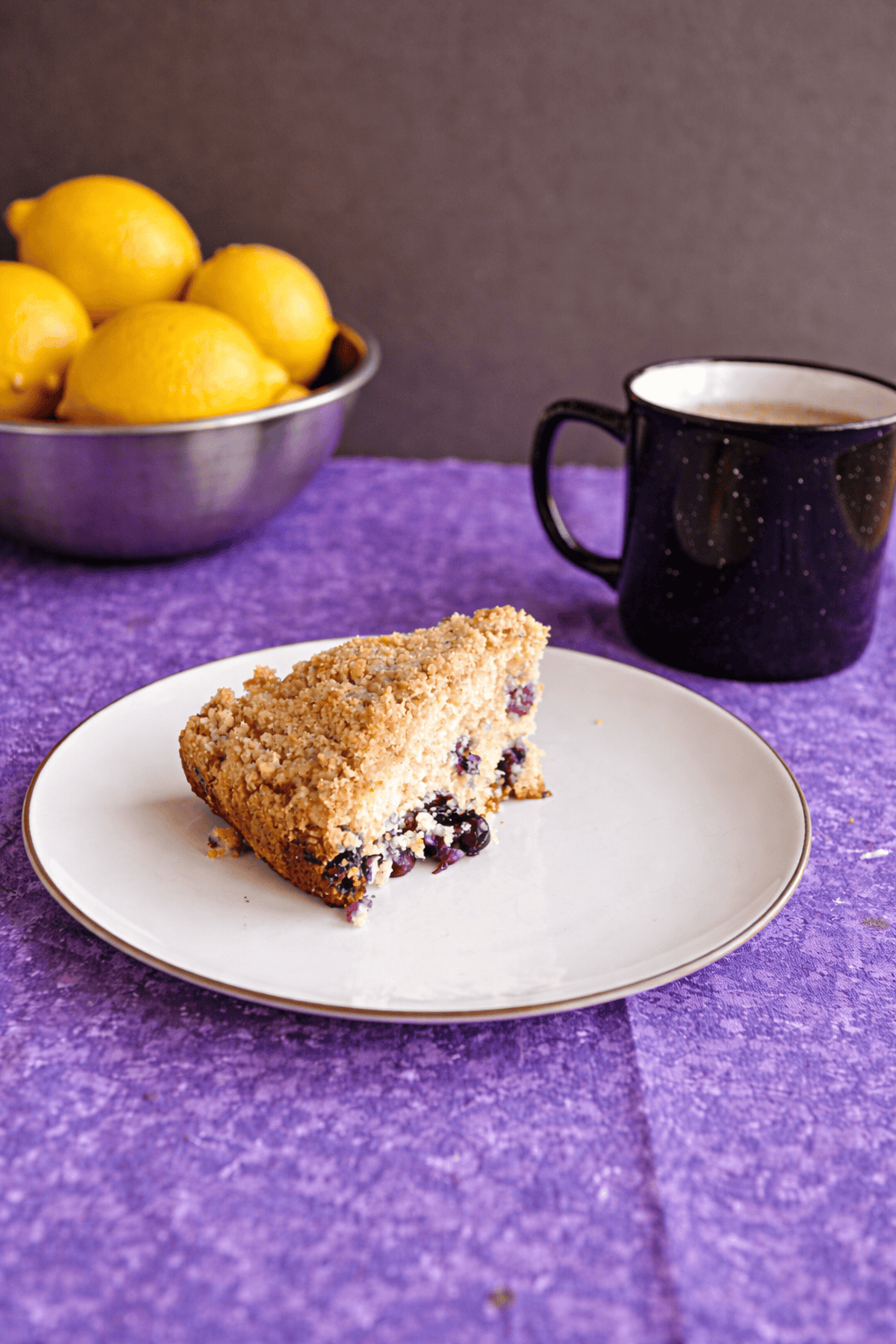 A plate with a slice of coffee cake on it, a mug of coffee behind the plate, and a bowl of lemons.