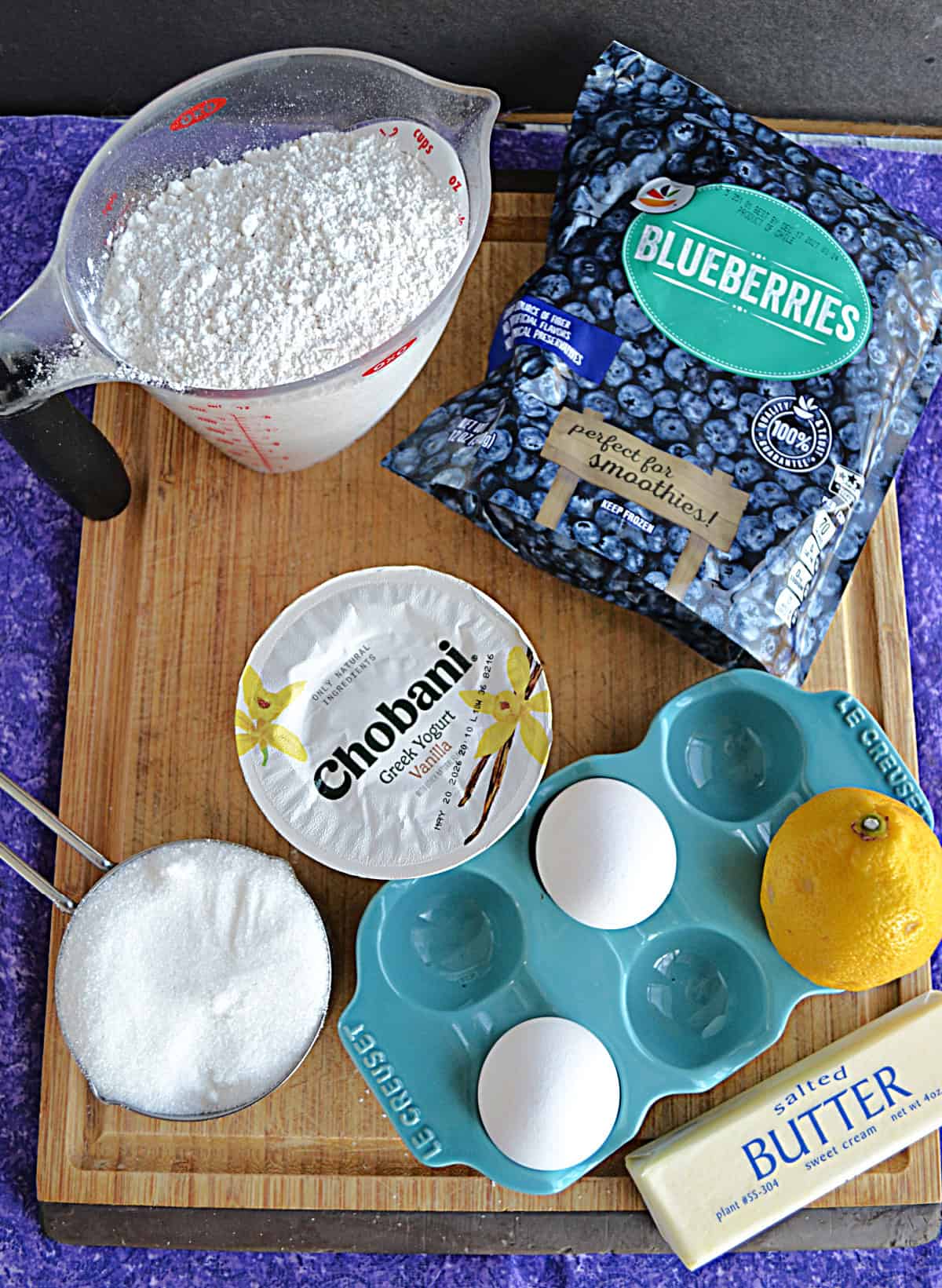 A cutting board with a cup of flour, a bag of frozen blueberries, a cup of yogurt, a lemon, 2 eggs, and a cup of sugar on it.