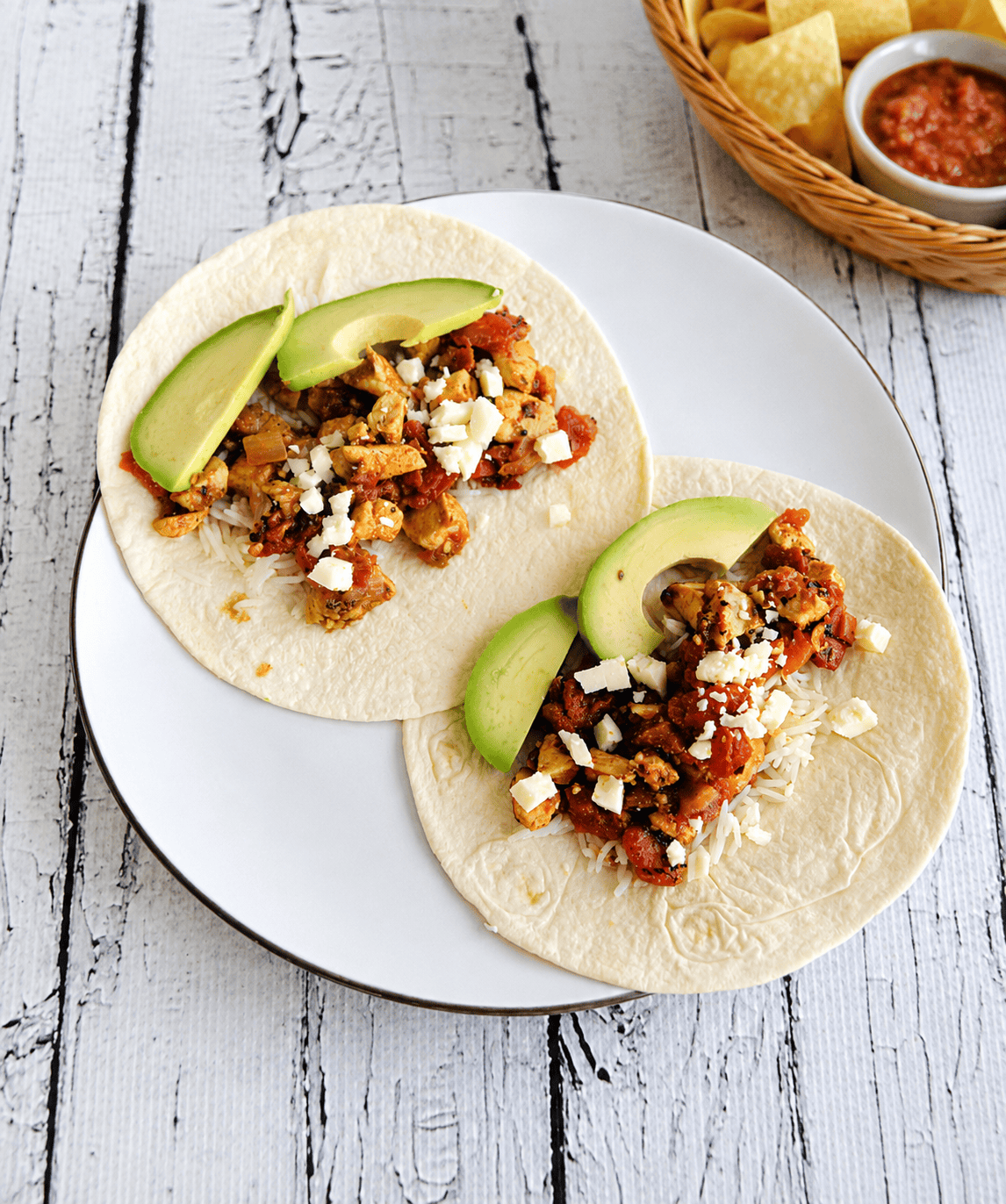 A plate with two chicken tacos on it and a bowl of chips and salsa behind the plate.
