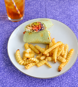 A plate with a green wrap cut in half and fries on it and a glass of iced tea in the background.