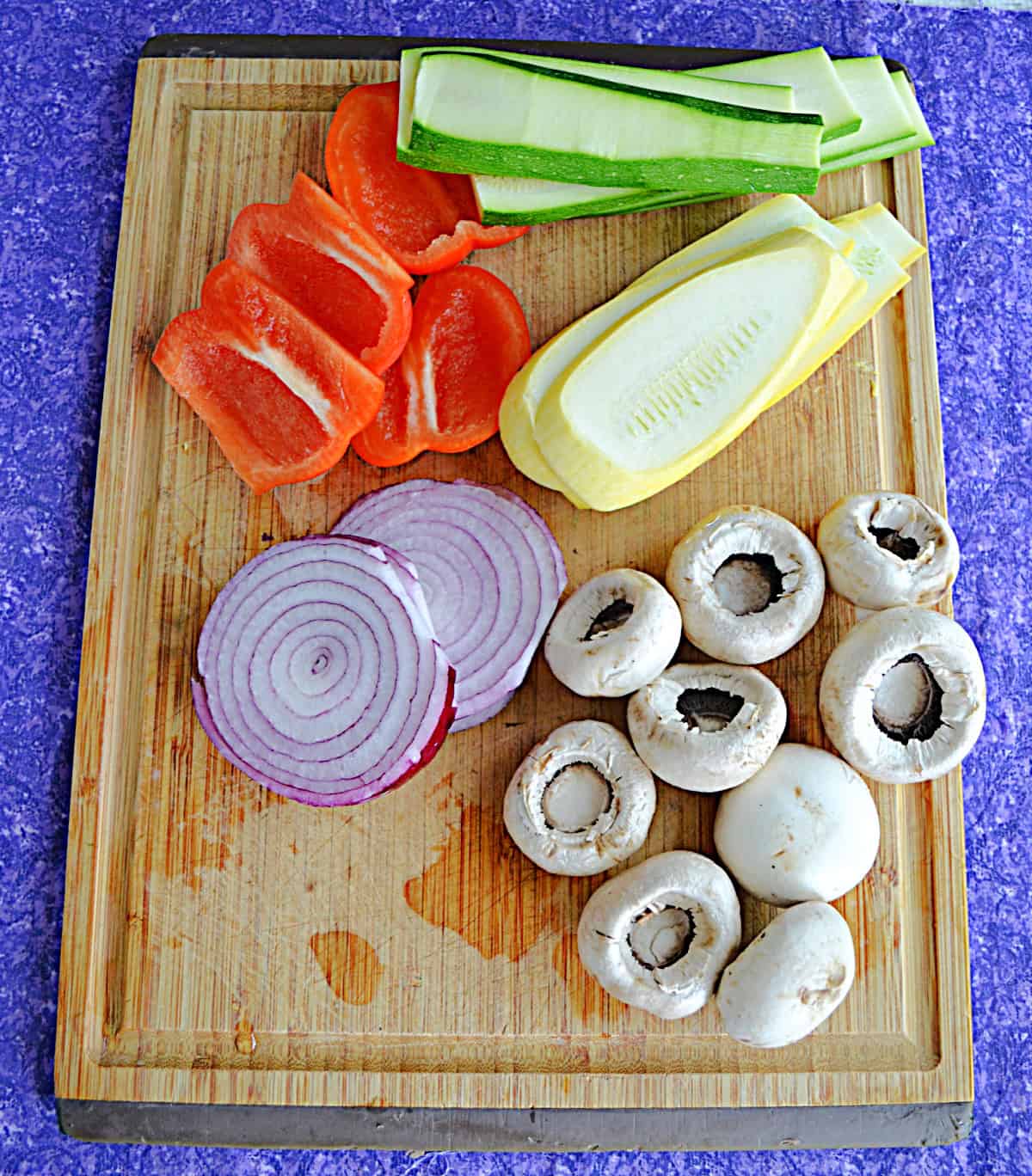 A wooden cutting board with sliced zucchini, squash, onions, red peppers, and mushrooms on it.