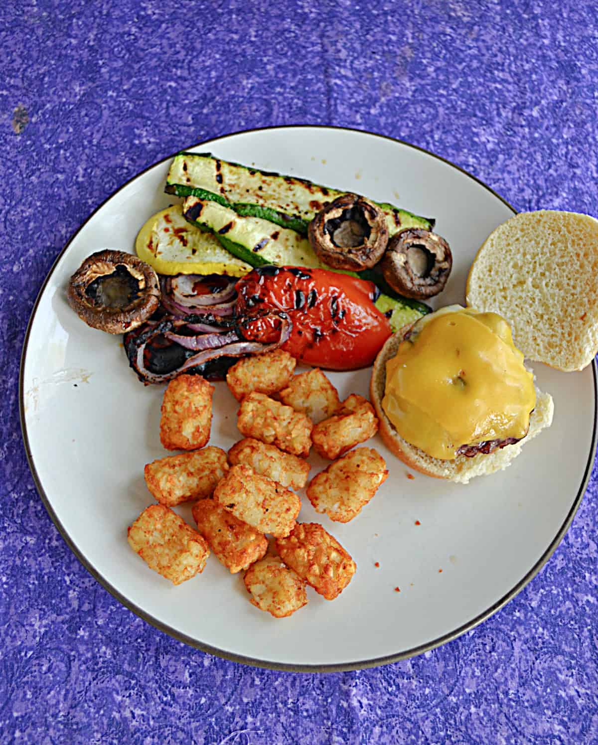A plate of grilled veggies, tater tots, and a cheeseburger.