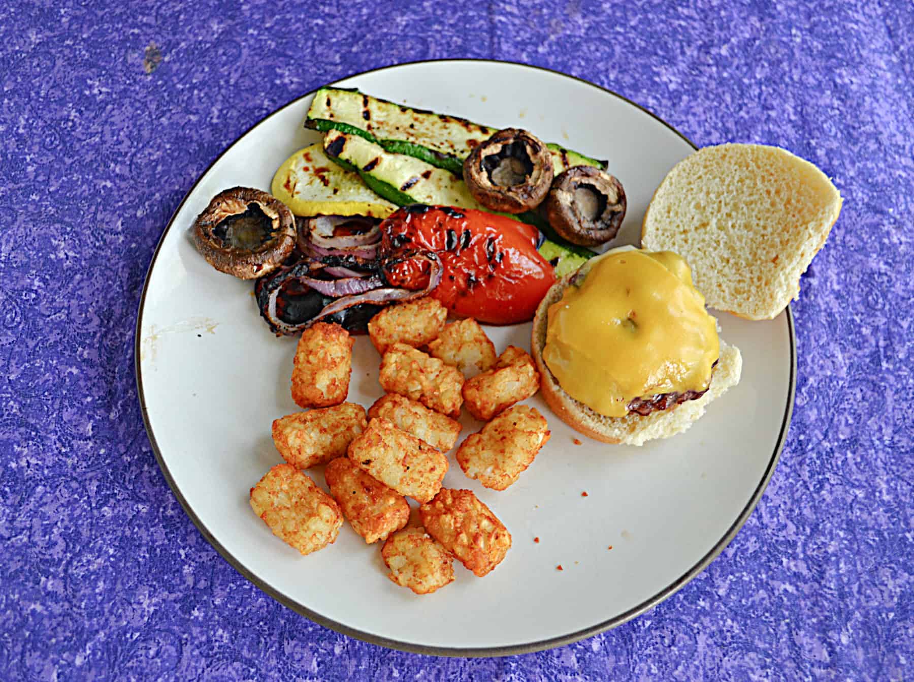 A plate with a variety of grilled vegetables, tater tots, and grilled cheeseburg.