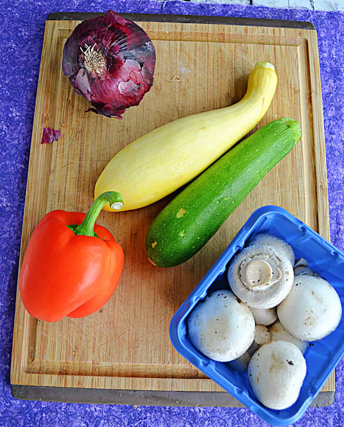 A wooden cutting board with whole zucchini, squash, onions, red peppers, and mushrooms on it.