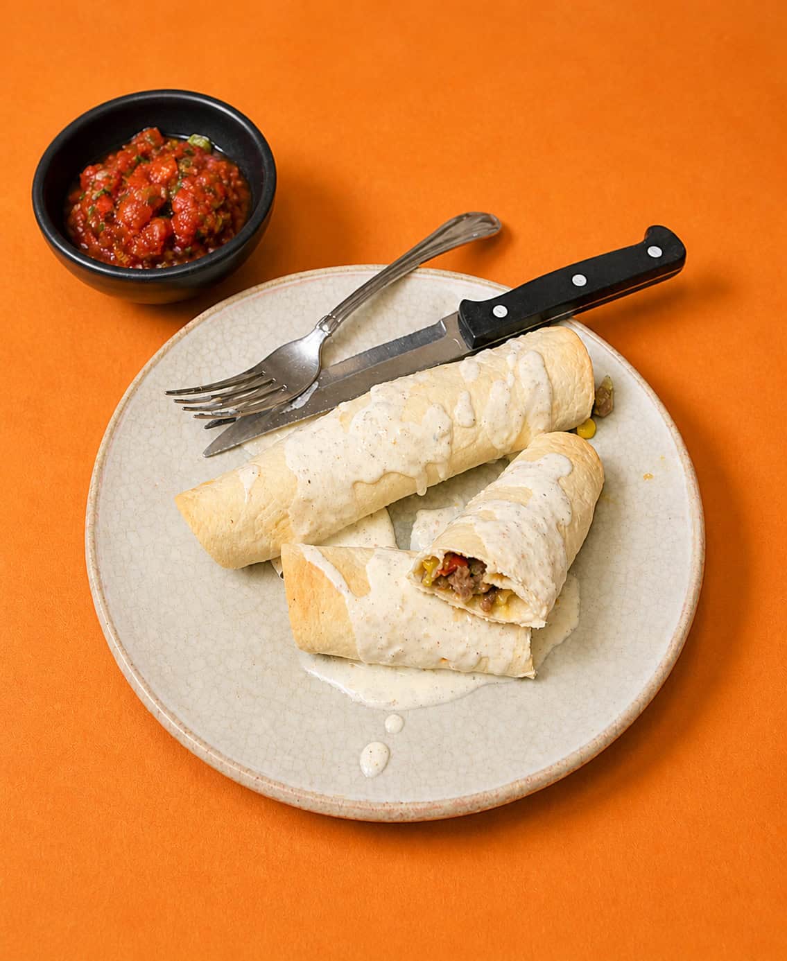 A plate of taquitos with a fork and knife on the plate and a bowl of salsa behind the plate.