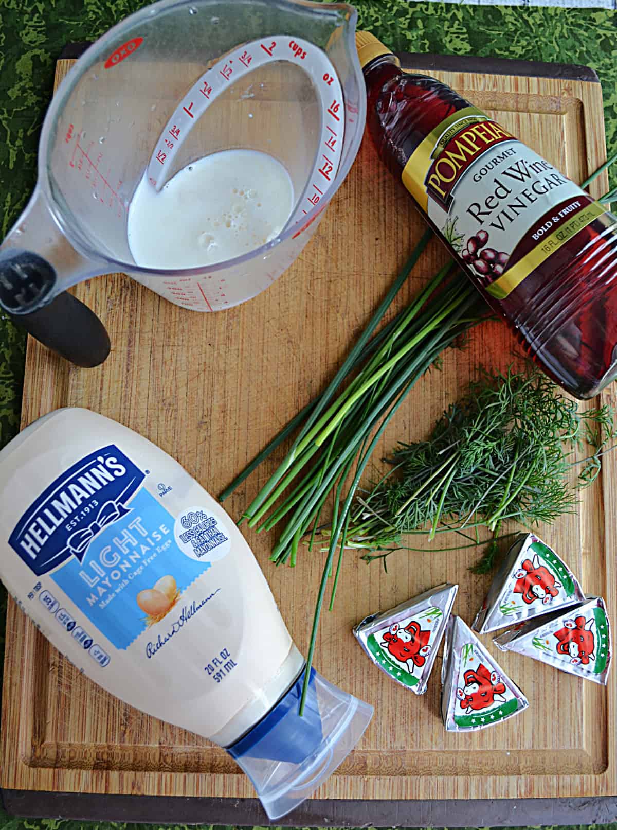 A cutting board with mayonnaise, fresh herbs, cheese, vinegar, and milk on it.