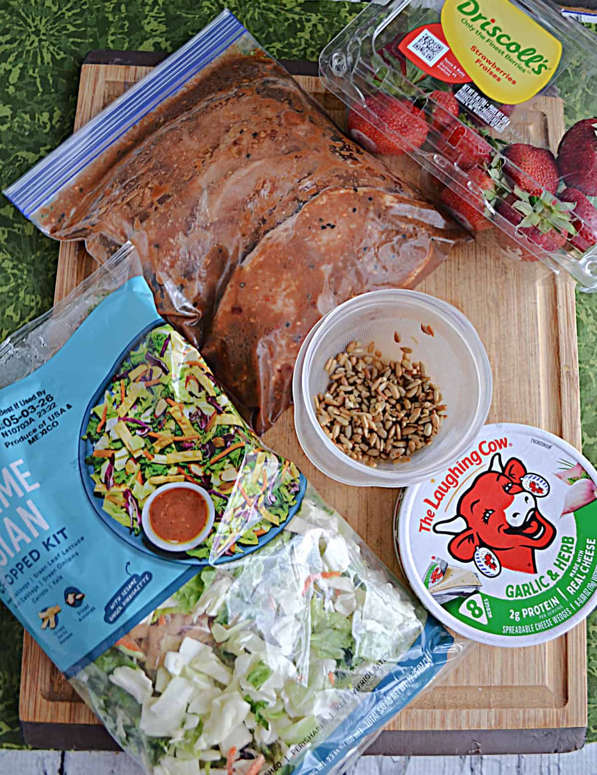 A cutting board with a bag of salad mix, cheese, marinated pork, a container of strawberries, and sesame seeds on it.