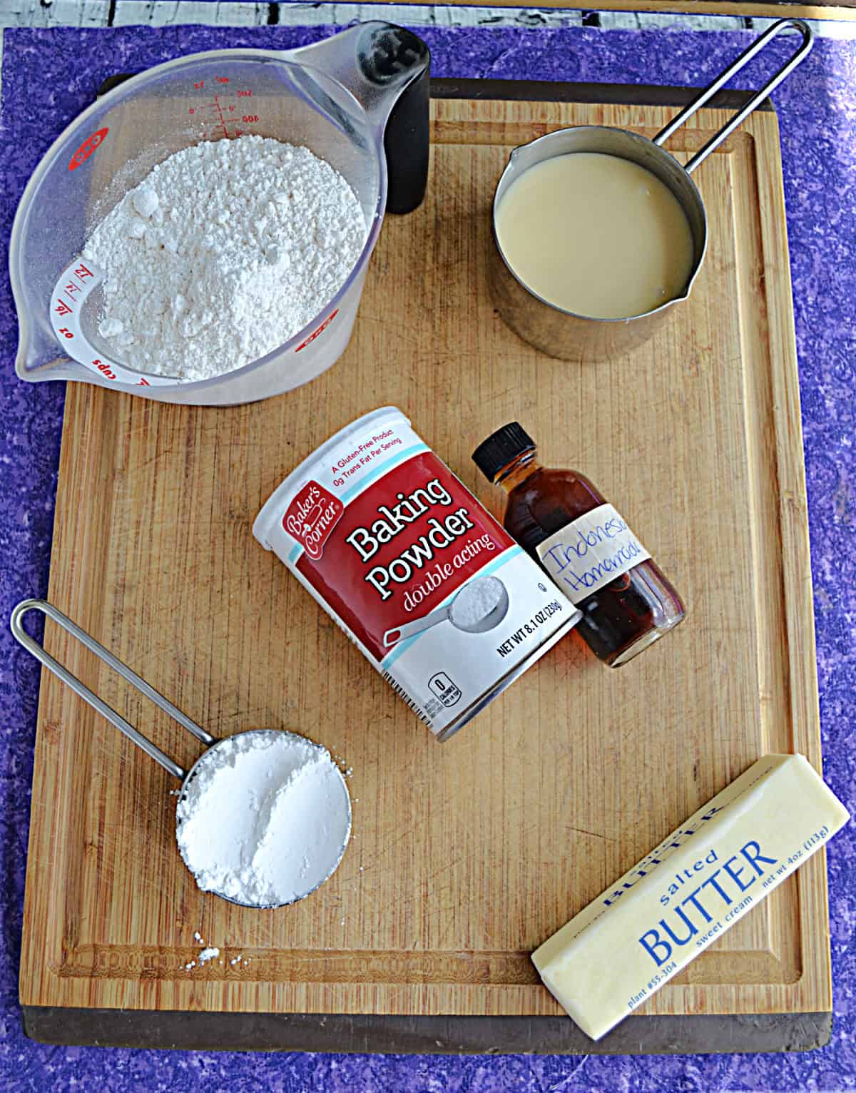 Wooden cutting board with a cup of flour, a stick of butter, a cup of condensed milk, vanilla, and baking powder on it.