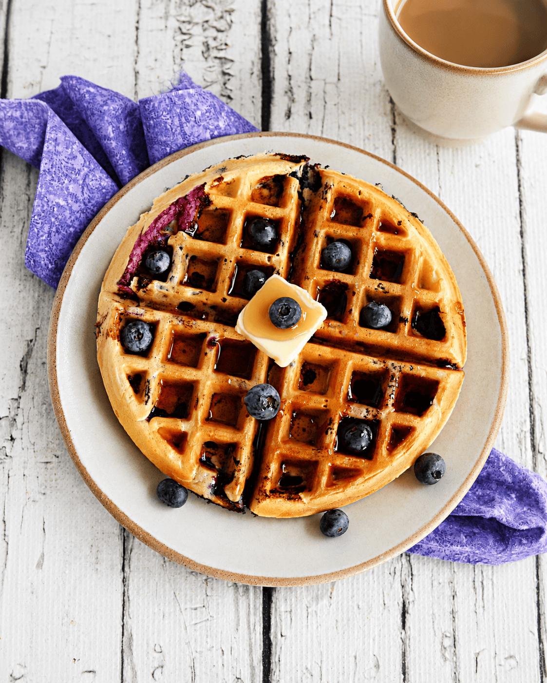 A waffle with fresh blueberries, butter, and syrup on it with a cup of coffee in the back.