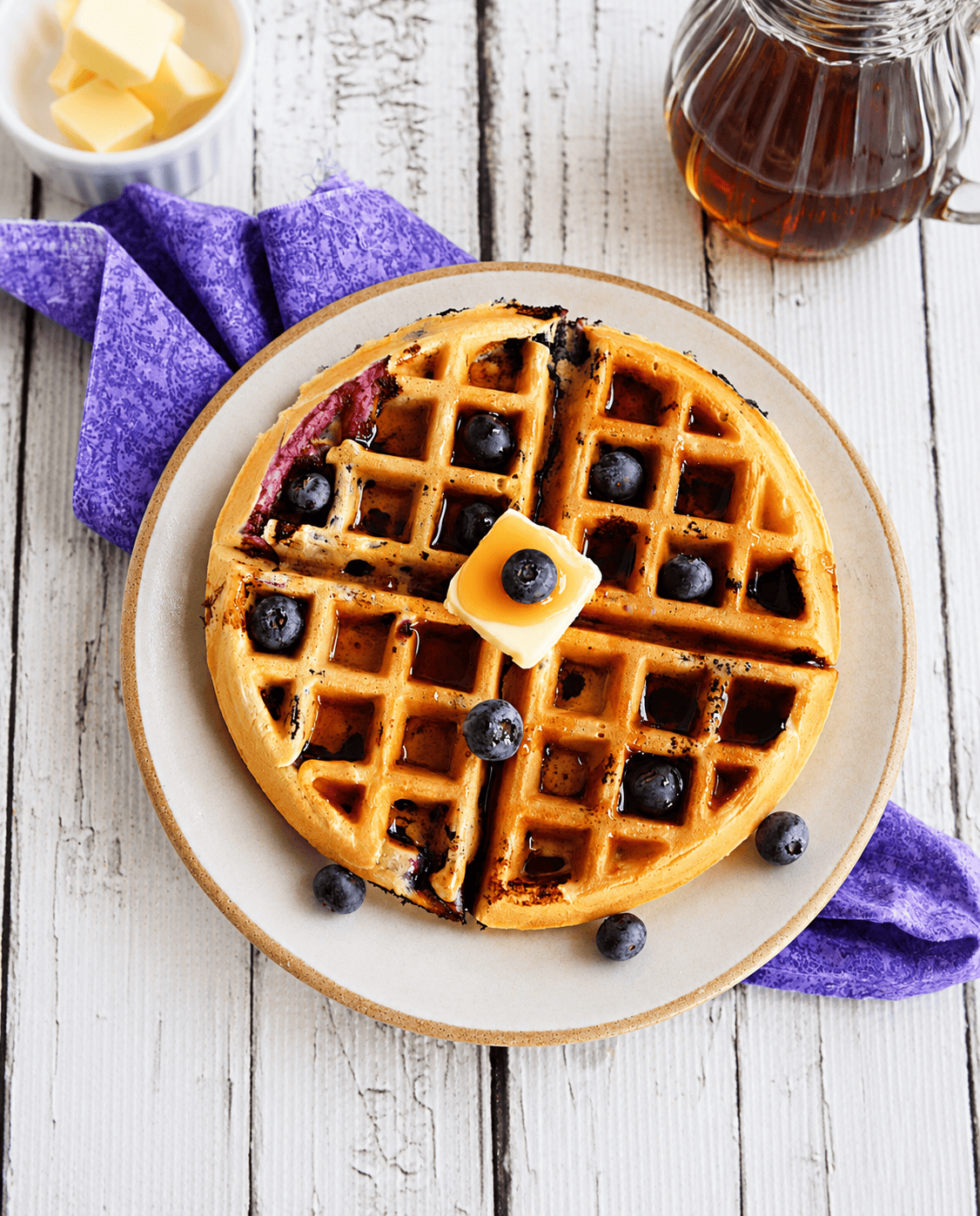 A waffle with fresh blueberries, butter, and syrup on it, a bowl of butter, and a carafe of syrup.