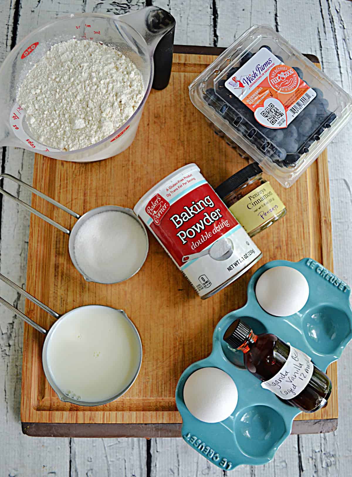 A cutting board with a cup of flour, cup of sugar, eggs, a container of blueberries, and cinnamon on it.