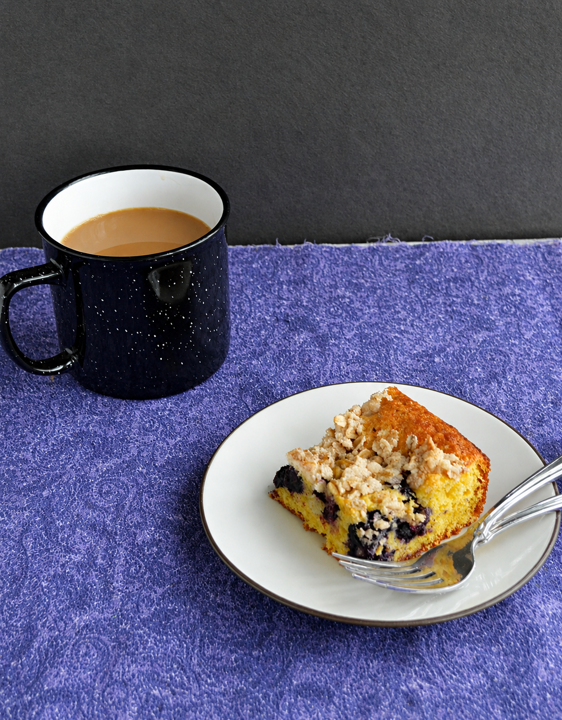 A plate with a piece of berry coffee cake and a fork on it with a cup of coffee in the background.