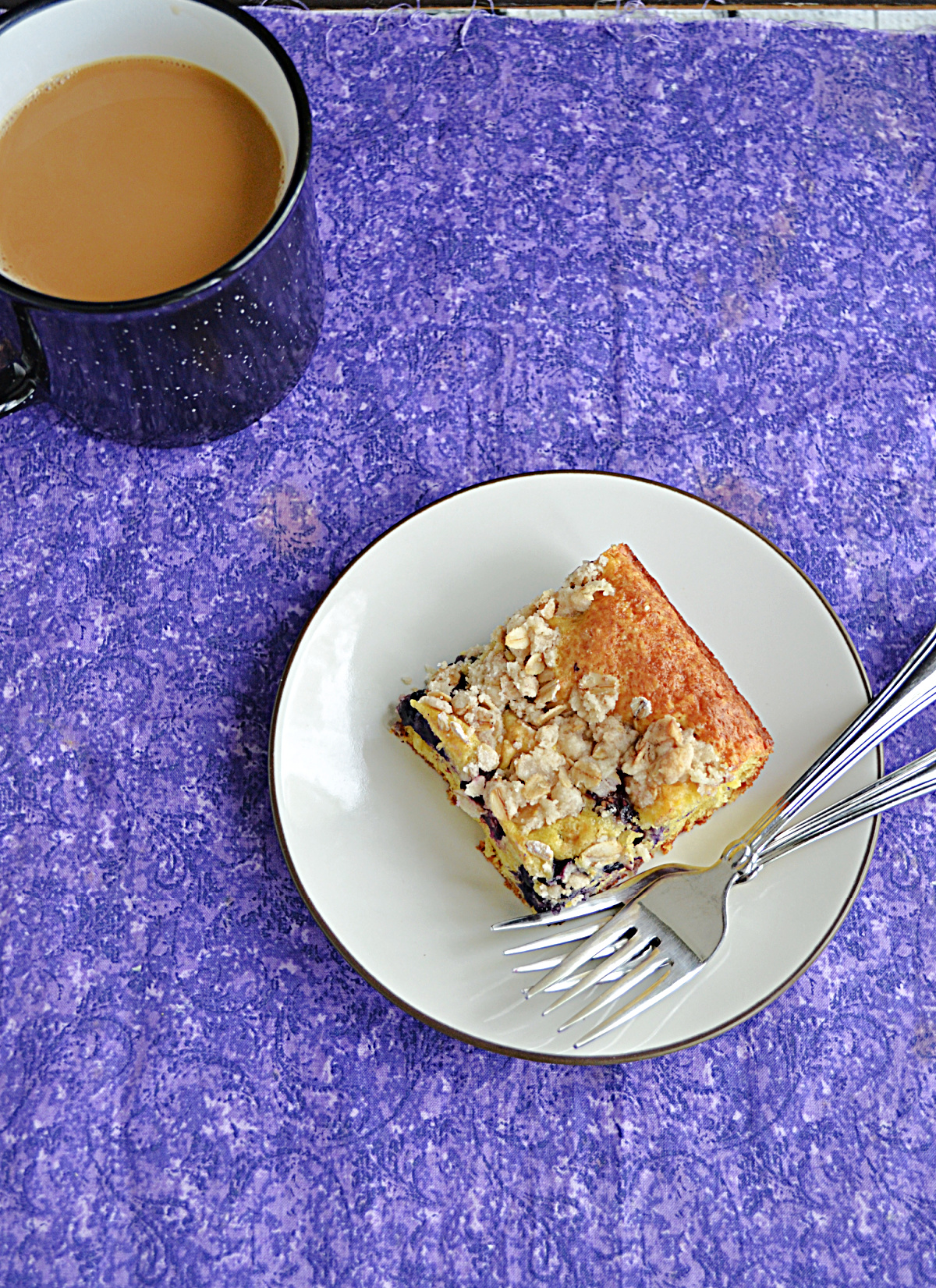 A top view of A plate with a piece of berry coffee cake and a fork on it with a cup of coffee in the background.