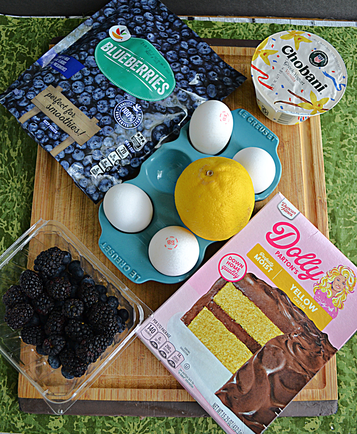 A cutting board with a box cake, blueberries, blackberries, 4 eggs, a lemon, and a cup of yogurt on it.