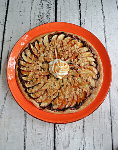 A plate with a dessert pizza with thinly sliced apples, a crumble, whipped cream, and a caramel drizzle.