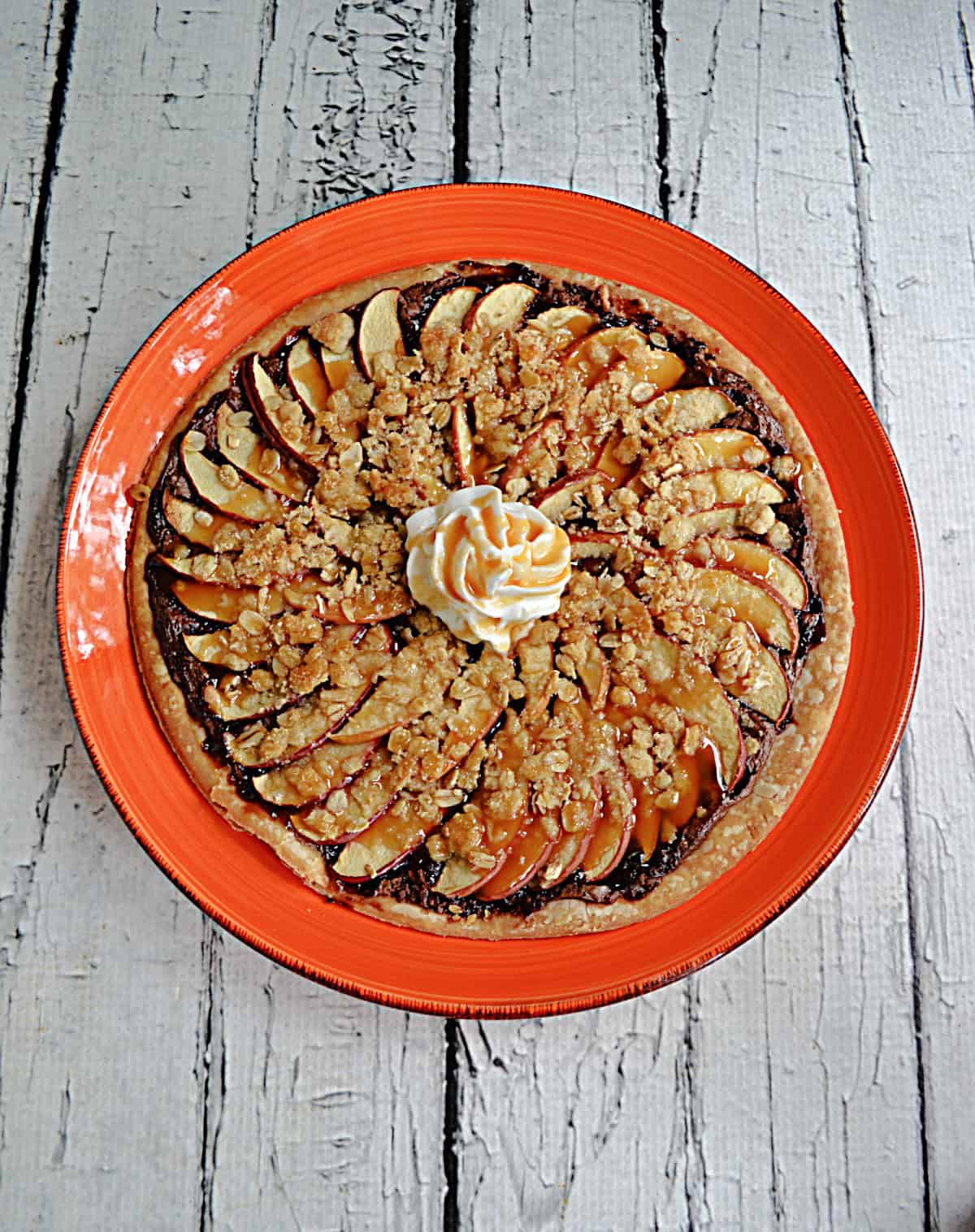 A plate with a dessert pizza with thinly sliced apples, a crumble, whipped cream, and a caramel drizzle.