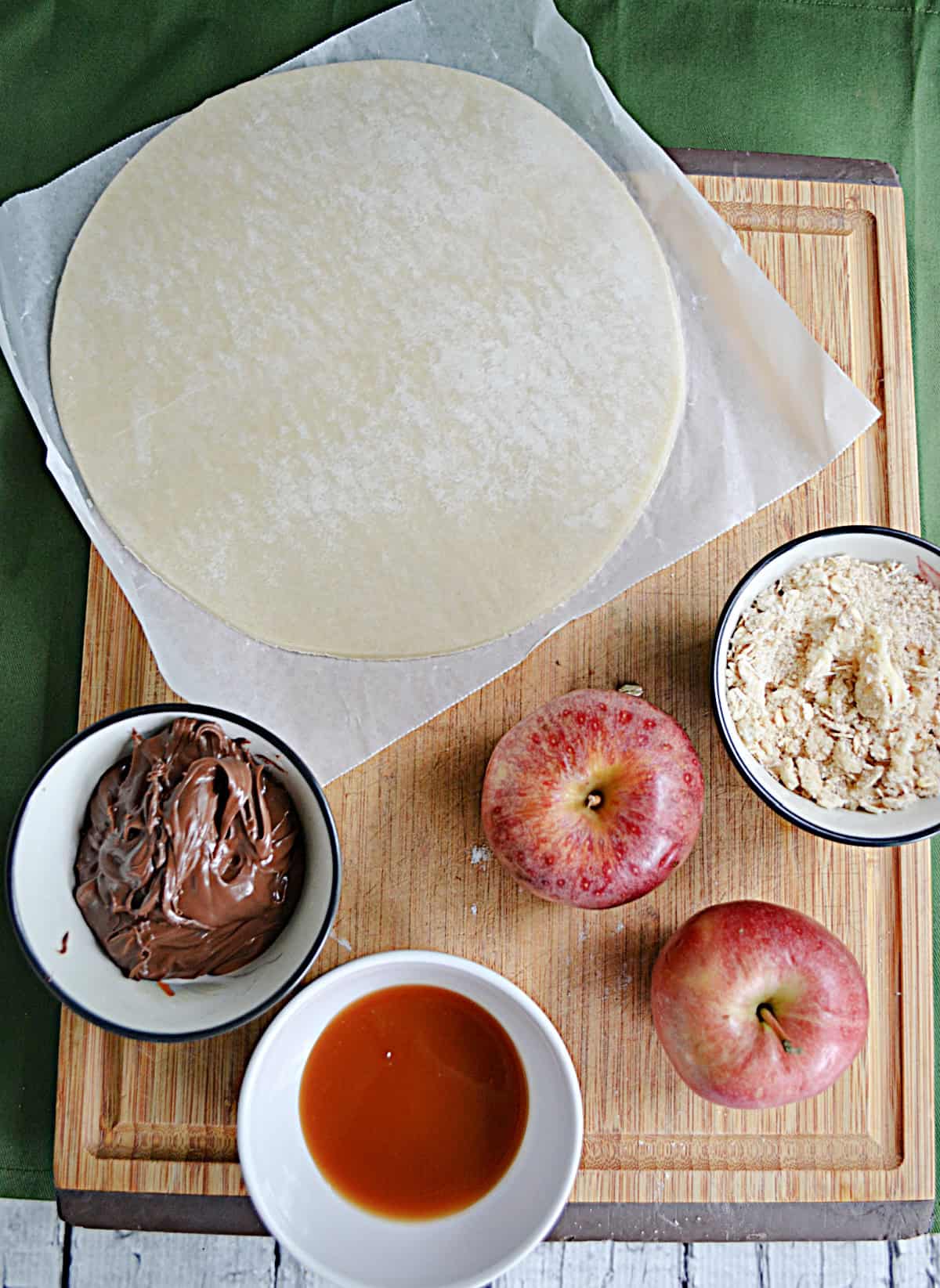A cutting board with a dough circle, apples, brown sugar, caramel, and chocolate hazelnut spread.