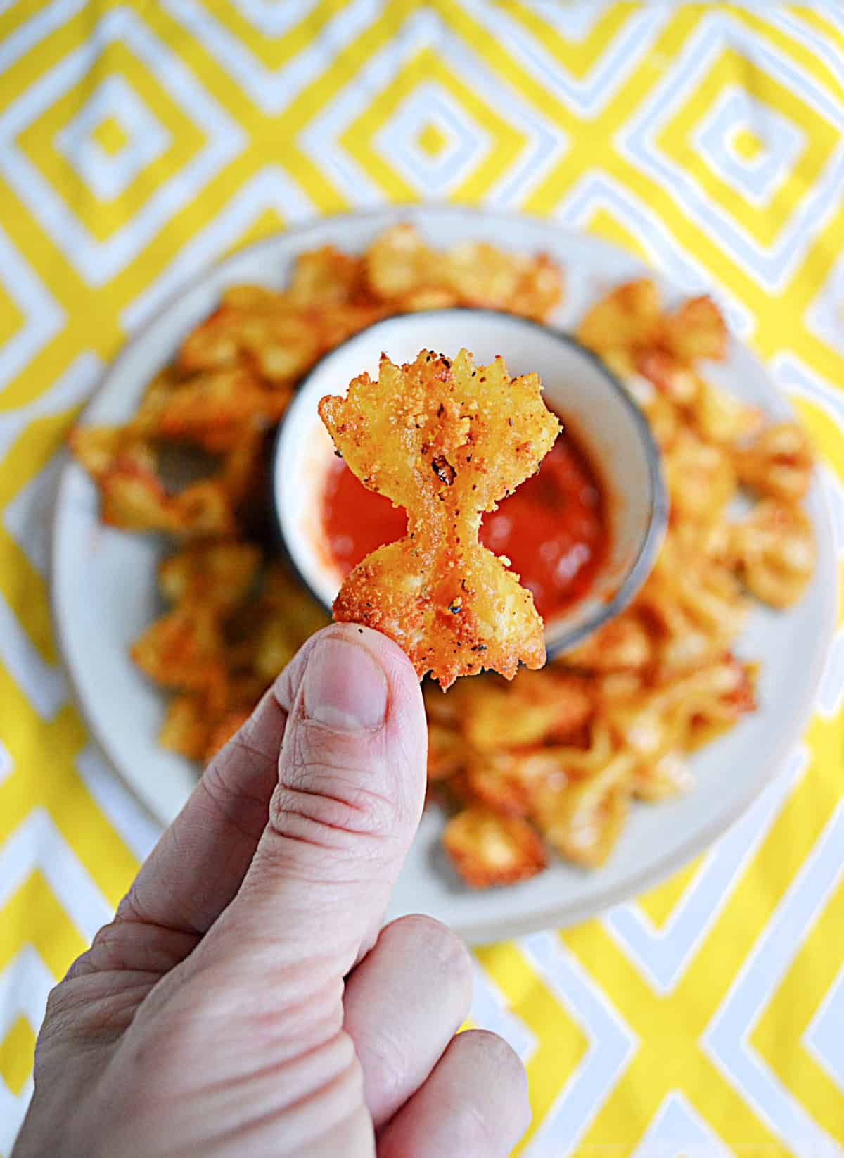 A hand holding a golden brown pasta chip over top of a plate of pasta chips.