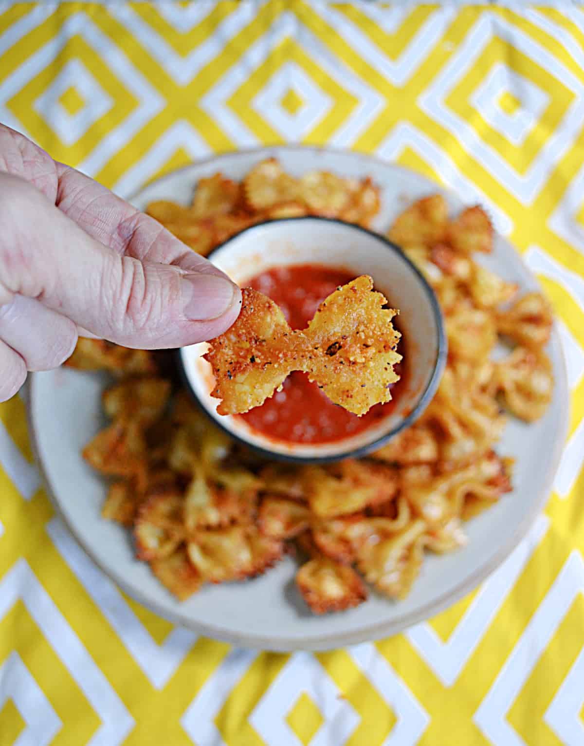 A hand holding a golden brown pasta chip over top of a bowl of marinara sauce.