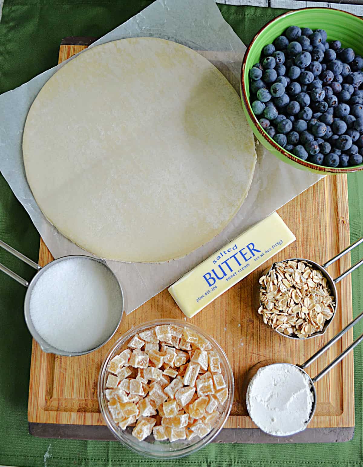 A cutting board with a dough cirlce, a bowl of blueberries, a stick of butter, a cup of sugar, a cup of oats, crystalized ginger, and cornstarch on it.