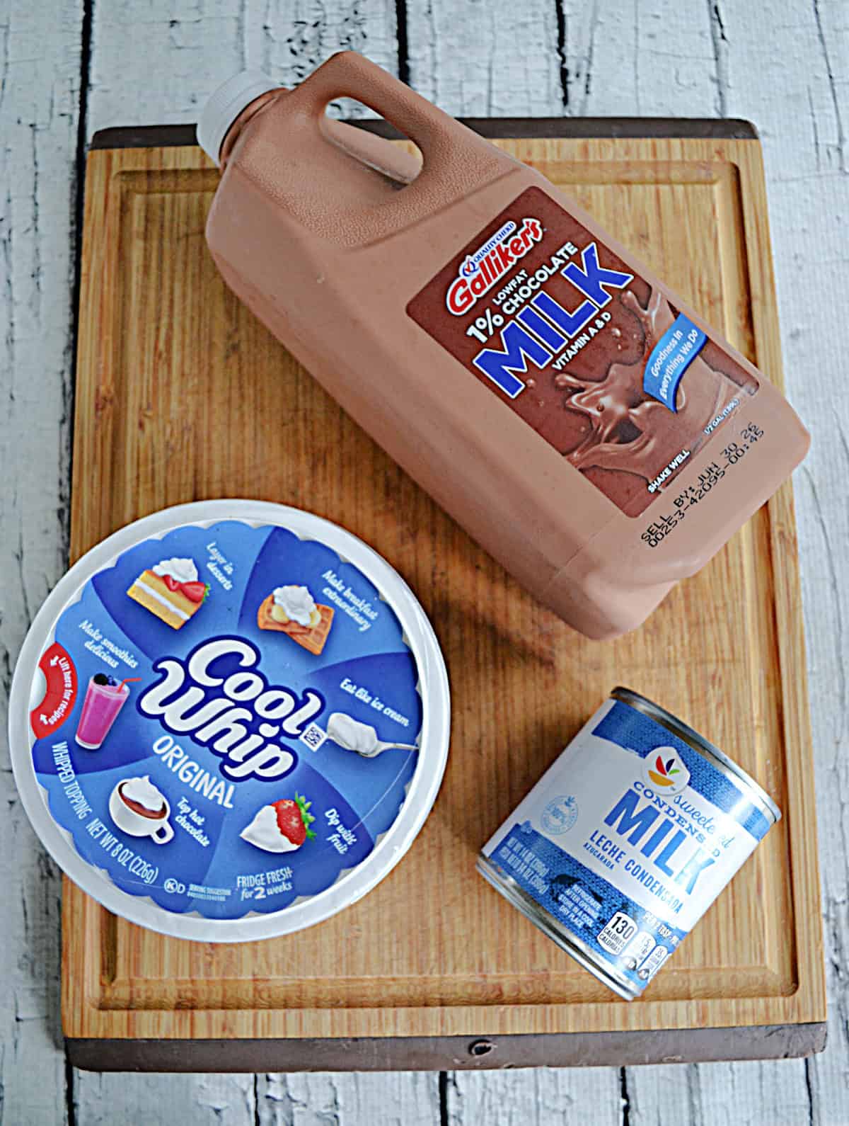 A cutting board with a gallon of chocolate milk, a container of Cool Whip, and a can of sweetened condensed milk on it.