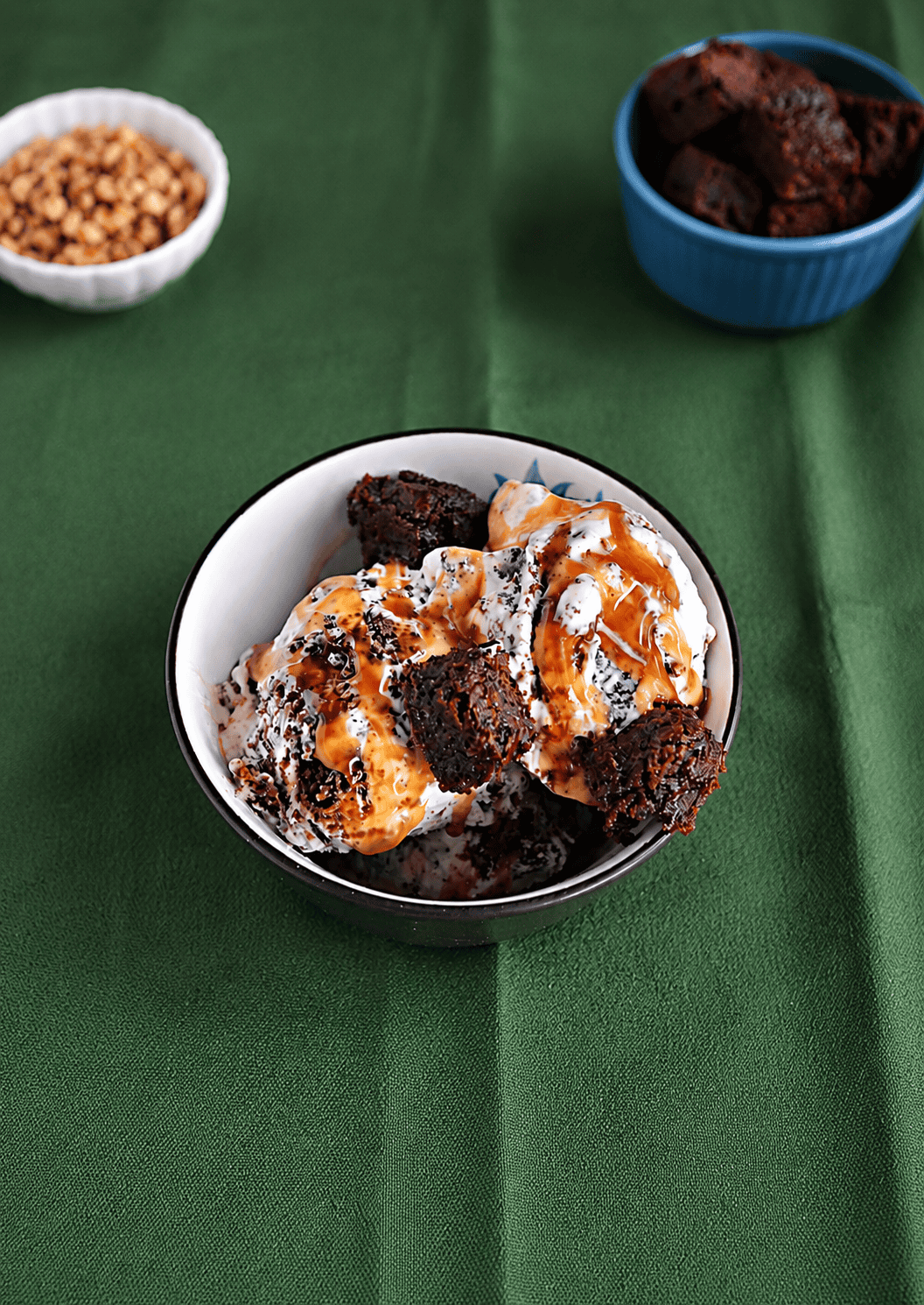 A bowl of caramel brownie ice cream with a bowl of brownie bites and a bowl of nuts behind it.