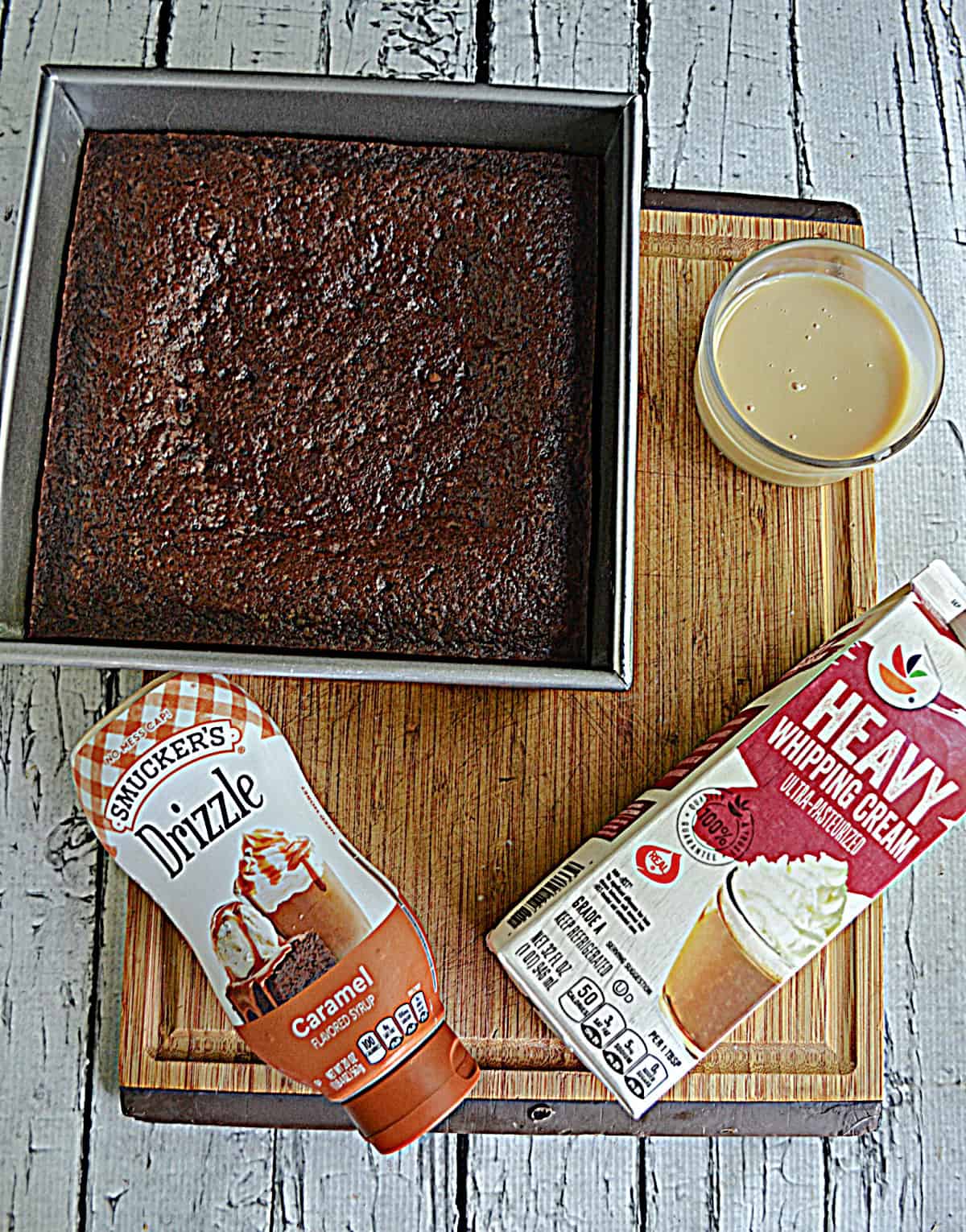 A pan of brownies, a bottle of caramel sauce, a quart of heavy cream, and a bowl of sweetened condensed milk.