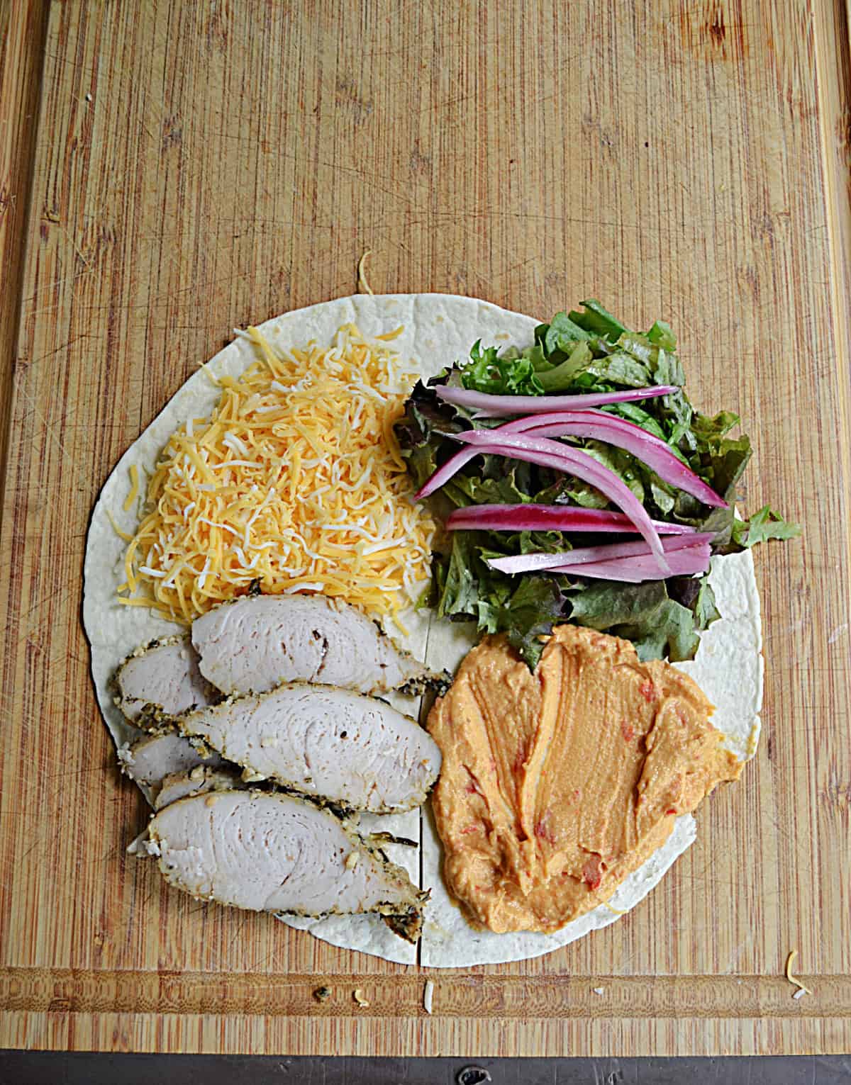 A tortilla with slices of turkey, cheese, hummus, and vegetables on it.